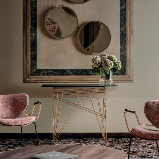 A contemporary interior features a Contemporary Glass Console Table with a geometric metal base and a dark tabletop, adorned with a vase of hydrangeas. Two pink fabric armchairs flank the table, while round, abstract mirrors with gold accents hang on the wall above.