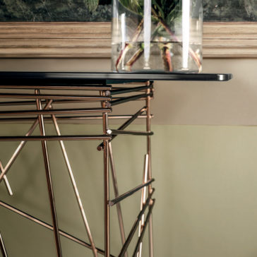 Contemporary Glass Console Table A close-up of the Contemporary Glass Console Table featuring a geometric wireframe base in metallic hues. Atop the table sits a transparent vase, showcasing plant stems inside. The backdrop includes framed artistic wall decor and a two-tone wall.