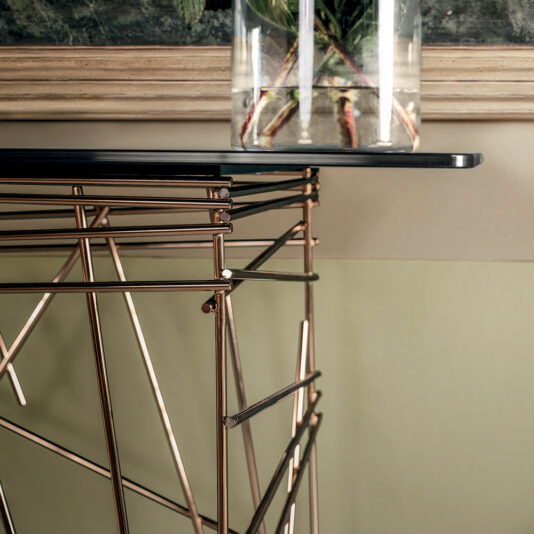 A close-up of the Contemporary Glass Console Table featuring a geometric wireframe base in metallic hues. Atop the table sits a transparent vase, showcasing plant stems inside. The backdrop includes framed artistic wall decor and a two-tone wall.