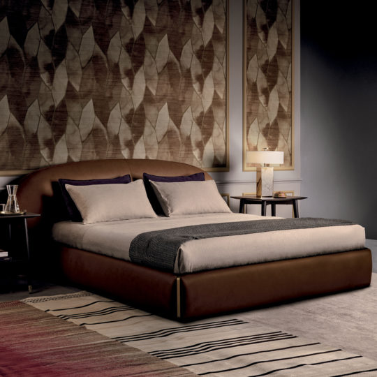 A contemporary bedroom featuring the Contemporary Leather Upholstered Curved Bed, elegantly dressed in beige and gray bedding. It is flanked by two round wooden nightstands, each topped with a lamp. The walls are adorned with large framed botanical artwork, and a striped rug lies beneath the bed on the floor.