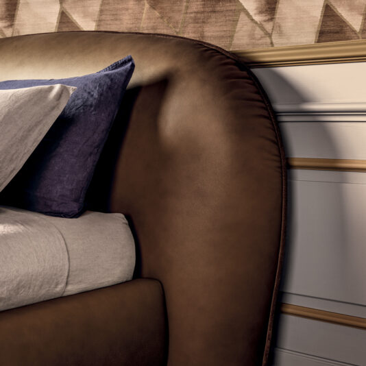 A close-up of a contemporary bedroom highlights the Contemporary Leather Upholstered Curved Bed. The dark brown leather bed frame is elegantly dressed with light gray sheets, complemented by both gray and purple pillows. The bed’s curved headboard adds a touch of sophistication, while geometrically patterned wallpaper can be seen partially in the background.