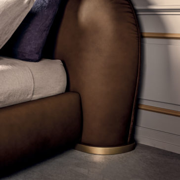 Contemporary Leather Upholstered Curved Bed A close-up view of the Contemporary Leather Upholstered Curved Bed showcases its luxurious design, featuring a rich brown headboard with a golden base. The bed, set against a white wall with decorative molding, partially displays a beige sheet and a purple pillow.