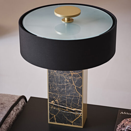Contemporary Marble Table Lamp The Contemporary Marble Table Lamp features a black cylindrical lampshade with a transparent glass top and a golden knob. Its rectangular base, adorned with black marble and gold veining, rests elegantly on a dark surface next to a partially visible book.