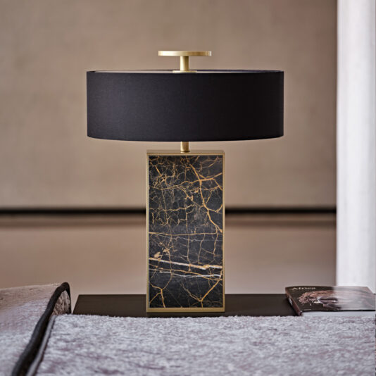 Contemporary Marble Table Lamp A Contemporary Marble Table Lamp with a rectangular base showcasing a black and gold marble pattern is complemented by its cylindrical black lampshade. The lamp sits on a dark tabletop beside an open book in a minimalist room with a beige background.