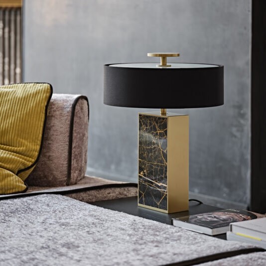 Contemporary Marble Table Lamp The Contemporary Marble Table Lamp, featuring a black rectangular shade and an elegantly patterned black and gold marble cylindrical base, sits on a dark side table. Positioned beside a stack of books, the lamp's sophisticated design effortlessly complements the yellow accent pillow on the gray textured sofa.