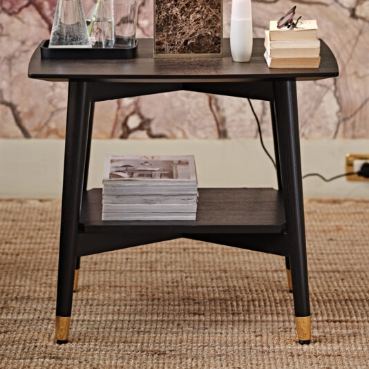 Contemporary Mocha Ash Bedside Table A Contemporary Mocha Ash Bedside Table with gold-tipped legs stands on a carpeted floor. The tabletop features a few stacked books, a pair of glasses, a small marble slab, and a water carafe set. The lower shelf holds a stack of magazines. In the background, there's a marbled wall.