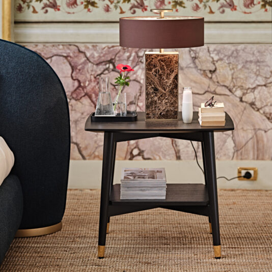 Contemporary Mocha Ash Bedside Table A Contemporary Mocha Ash Bedside Table with a two-tiered black surface, featuring a brown marble lamp, a vase holding a red flower, a glass water carafe, a white gadget, and stacks of books and magazines. The background showcases detailed wallpaper in pink and beige tones.