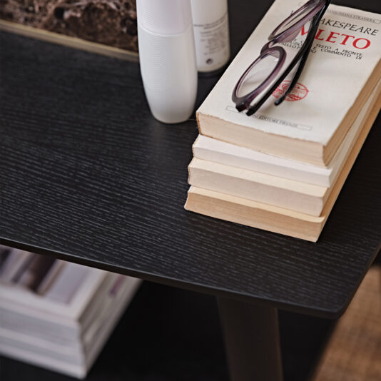 Contemporary Mocha Ash Bedside Table A close-up of a black wooden side table, almost reminiscent of the Contemporary Mocha Ash Bedside Table, features a stack of paperback books topped with glasses and a couple of white bottles. One book titled