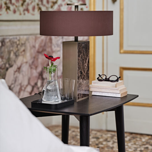 Contemporary Mocha Ash Bedside Table A Contemporary Mocha Ash Bedside Table with a sleek black top supports a chic brown lamp, a glass vase containing a vibrant red flower, an empty glass, a stack of books, and a pair of eyeglasses. The scene is set against a decorative wall and an elegant door with gold trim.