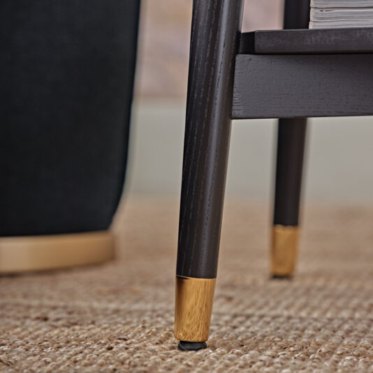 Contemporary Mocha Ash Bedside Table Close-up of a dark wooden furniture leg with a gold-colored metal tip, resting on a beige textured rug. The background includes a partial view of another dark-colored furniture piece with a gold base, perfectly complementing the Contemporary Mocha Ash Bedside Table.