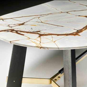 A close-up of the Contemporary Oval Quartz Dining Table reveals its elegant, white surface adorned with intricate golden-tinged cracks. This refined tabletop is supported by dark wooden legs and brass-colored metallic crossbars, offering a striking contrast between materials.
