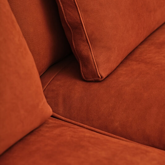 Close-up of an orange-red suede couch with matching cushions. The image highlights the texture of the suede material and the finely stitched seams of the cushions against this Contemporary Retro Inspired Leather Sofa.