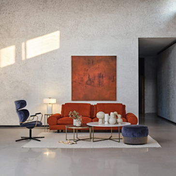 Contemporary Retro Inspired Leather Sofa A modern living room features a Contemporary Retro Inspired Leather Sofa adorned with multiple cushions, a coffee table with decorative items, a side table with a lamp, and a blue armchair. There's a large abstract painting on the wall behind the sofa, and sunlight streams through an unseen window.