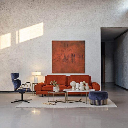 A modern living room features a Contemporary Retro Inspired Leather Sofa adorned with multiple cushions, a coffee table with decorative items, a side table with a lamp, and a blue armchair. There's a large abstract painting on the wall behind the sofa, and sunlight streams through an unseen window.