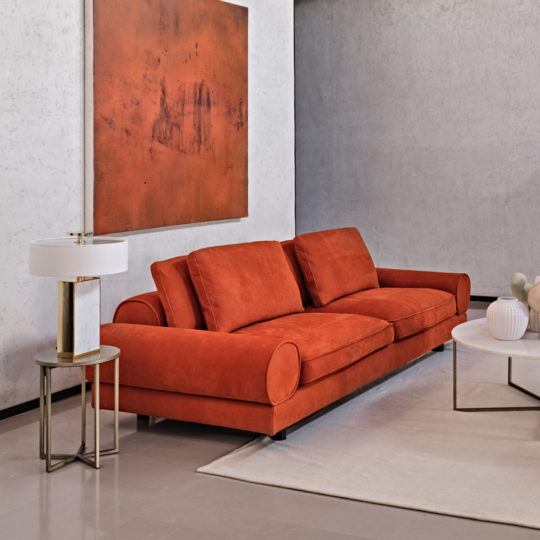 A modern living room features a Contemporary Retro Inspired Leather Sofa with cylindrical pillows against a grey wall. Above the sofa hangs an abstract painting in shades of orange. A white lamp sits on a small round side table next to the sofa, and a white rug is on the floor.