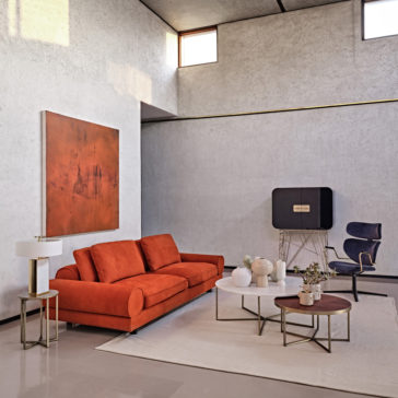 Contemporary Retro Inspired Leather Sofa A modern living room with high ceilings, grey walls, and tall windows showcases a Contemporary Retro Inspired Leather Sofa in red. The space is complemented by a square gold and white lamp, a round white coffee table adorned with decorative items, a smaller wooden table, and a black chair next to a unique sideboard in the corner.