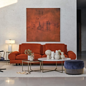 Contemporary Retro Inspired Leather Sofa A modern living room features a Contemporary Retro Inspired Leather Sofa in burnt orange with matching artwork above it. In front of the sofa are three round coffee tables in varying heights, decorated with white vases and a small potted plant. A blue pouf is placed next to the tables, and a floor lamp stands to the left, giving off contemporary retro vibes.