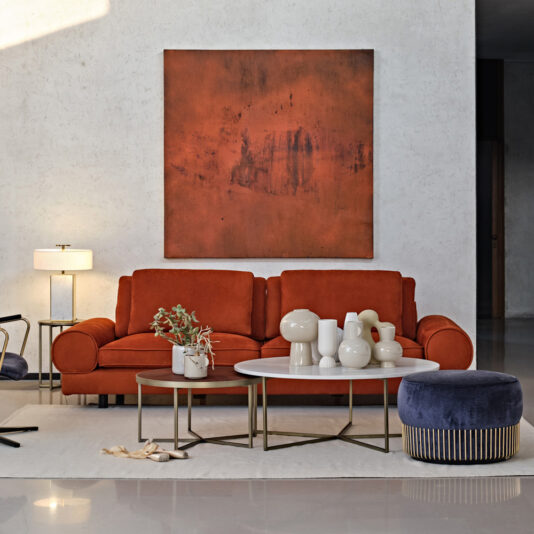 A modern living room features a Contemporary Retro Inspired Leather Sofa in burnt orange with matching artwork above it. In front of the sofa are three round coffee tables in varying heights, decorated with white vases and a small potted plant. A blue pouf is placed next to the tables, and a floor lamp stands to the left, giving off contemporary retro vibes.