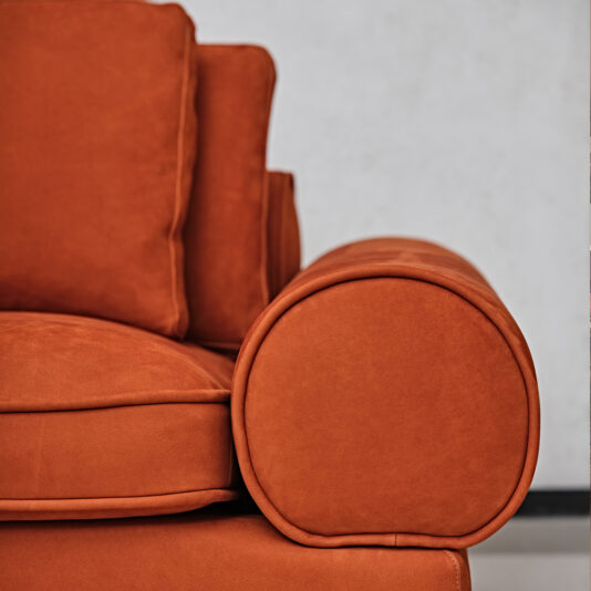 A close-up shot of the armrest of a Contemporary Retro Inspired Leather Sofa. The round and plush armrest adds to the sofa's inviting appearance, showcasing its soft and comfortable cushion. The plain, light-colored wall in the background contrasts beautifully with the sofa's vibrant color.
