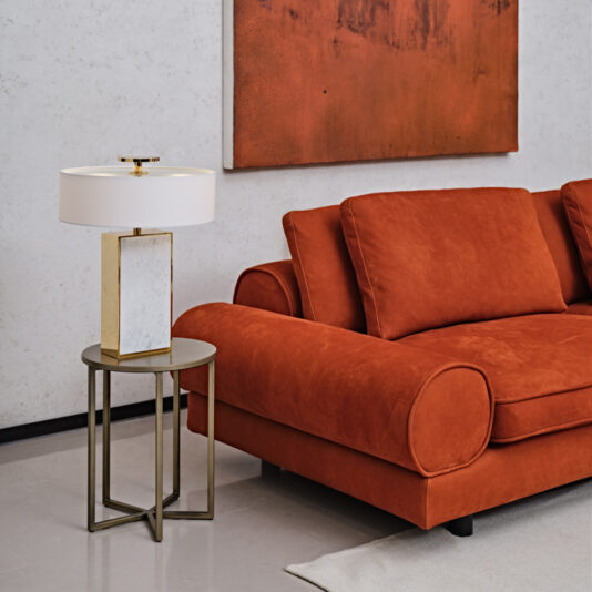 A contemporary living room showcases a Contemporary Retro Inspired Leather Sofa in burnt orange with roll arms, an abstract art piece on the wall, and a small round side table. The table holds a rectangular base lamp with a white shade and gold accents. The floor and walls are light-colored.