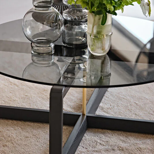 Contemporary Low Round Glass Coffee Table The Contemporary Low Round Glass Coffee Table with its black metal frame is adorned with various decorative items, including clear glass vases and a potted plant with green foliage. The table stands elegantly on a beige carpet, adding a touch of modern sophistication to the room.