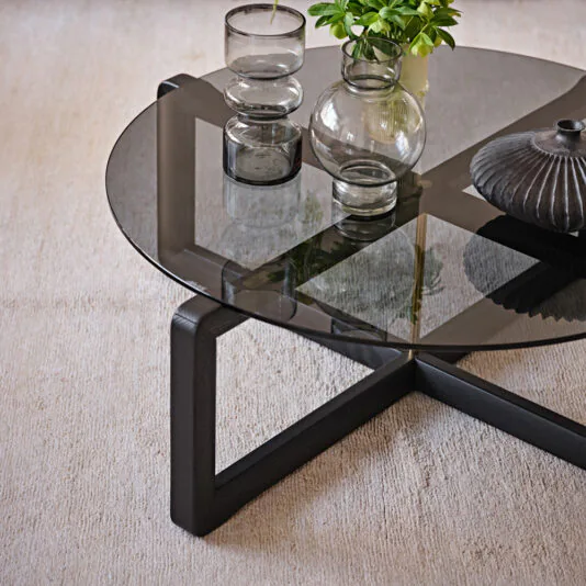 Contemporary Low Round Glass Coffee Table The product, the Contemporary Low Round Glass Coffee Table, featuring a black wooden frame, is positioned on a light-colored carpet. It is elegantly decorated with two modern glass vases: one small vase with a bubble design and another tapered vase that holds a leafy green plant, along with a textured black ceramic vase.