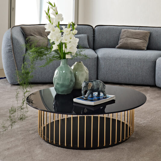 A modern living room showcases the Contemporary Round Glass Coffee Table, which features black accents and gold vertical stripes. The table is tastefully decorated with vases of white flowers and greenery, along with a small decorative elephant statue. Completing the setting are a gray couch and a gray rug.