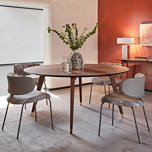 A contemporary dining area featuring the Contemporary Round Walnut Dining Table Set with four grey, cushioned chairs. The table is adorned with a glass vase holding green and white flowers. In the background, a side console shows a lamp and artwork against a burnt orange accent wall.