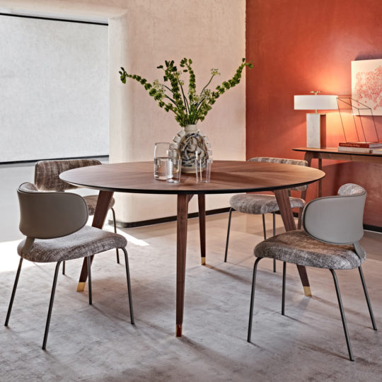 A contemporary dining area featuring the Contemporary Round Walnut Dining Table Set with four grey, cushioned chairs. The table is adorned with a glass vase holding green and white flowers. In the background, a side console shows a lamp and artwork against a burnt orange accent wall.
