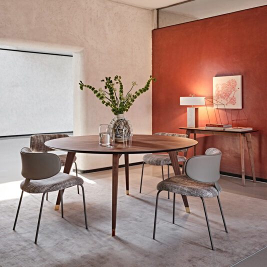A modern dining area featuring a Contemporary Round Walnut Dining Table Set adorned with a vase of flowers and a water pitcher. Four upholstered chairs encircle the table. The room boasts red and beige walls, a gray floor rug, and is accentuated by a side table holding a lamp and artwork.