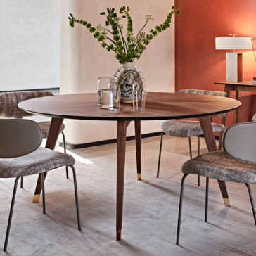 Contemporary Round Walnut Dining Table Set The Contemporary Round Walnut Dining Table Set, accompanied by four mid-century modern chairs, encircles a decorative vase filled with green foliage. Positioned on a light-colored rug with a red accent wall and a white lamp in the background, it creates a stylish dining space.