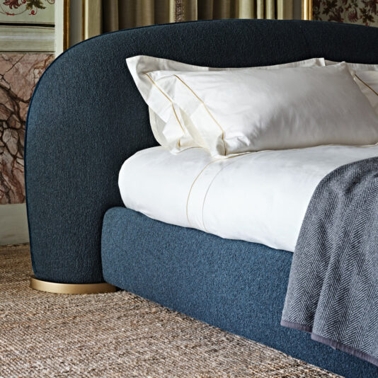 The Contemporary Upholstered Curved Bed features a sophisticated blue headboard and golden base, adorned with white pillows and a subtly patterned blanket, all set on a textured carpet.