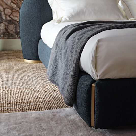 A sleek, modern _Contemporary Upholstered Curved Bed_ in dark blue with metallic accents sits atop a neutral woven rug. The bed is neatly made with white bedding and a dark gray herringbone-patterned throw draped over the side. The room's design is elegant, featuring textured wall details.