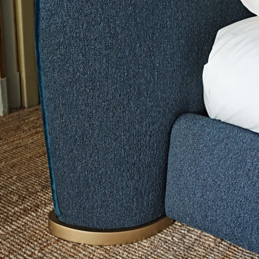Close-up of a Contemporary Upholstered Curved Bed featuring textured blue fabric. The elegant foot of the curved bed stands on a coarse woven rug. White bedding contrasts beautifully with the deep blue, while a gold accent at the base enhances its elegance.