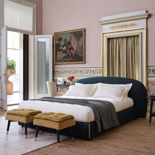 A sophisticated bedroom features the Contemporary Upholstered Curved Bed with white linens and a dark gray headboard. At the foot of the bed are two mustard-colored ottomans. The room is decorated with ornate moldings, a large classical painting, and an open balcony door that invites natural light.