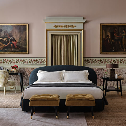 A luxurious bedroom features the Contemporary Upholstered Curved Bed with plush white linens, flanked by two brown cushioned benches. The room is adorned with classical paintings, ornate trim, floral wallpaper, and a desk paired with a wicker chair. A beige curtain hangs centrally behind the bed.