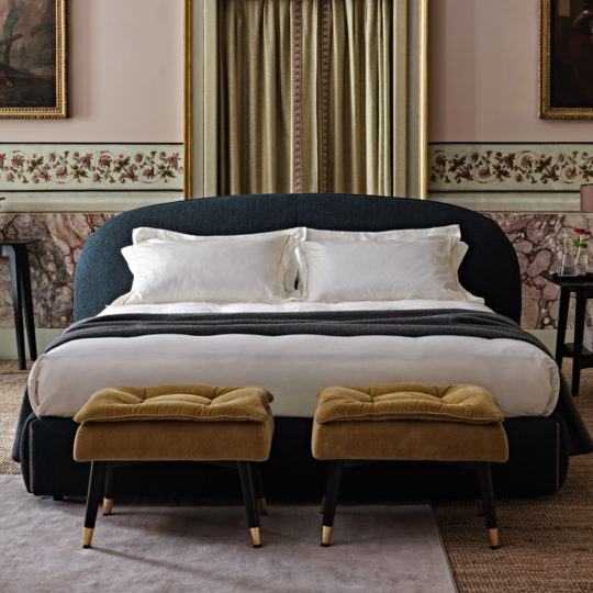 The luxurious bedroom boasts a Contemporary Upholstered Curved Bed adorned with white pillows and bedding. At its foot, two mustard-colored cushioned stools add a pop of color. The room's elegant atmosphere is heightened by ornate wall detailing, curtains, and framed artwork.