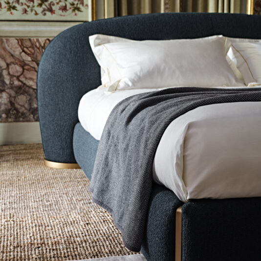The Contemporary Upholstered Curved Bed is showcased in a luxurious and modern setting with dark tones and golden accents. It features pristine white bedding and a neatly placed gray herringbone-patterned throw blanket. In the background, the room is adorned with a textured rug and intricate wall art, adding an extra touch of elegance.