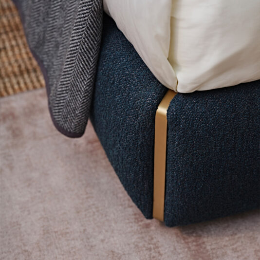 Close-up of the bottom corner of the Contemporary Upholstered Curved Bed, featuring a dark blue fabric-covered frame with a brass accent. The scene showcases part of a gray herringbone patterned blanket and white bedding, resting on a light pink carpet next to a section of a woven rug.