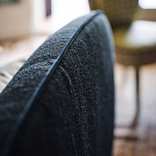 Close-up of the side of a dark gray, contemporary upholstered chair with textured fabric. The background is blurred, revealing part of a woven chair and wooden flooring. The image focuses on the chair’s quality and fabric details, evoking the craftsmanship and design of our Contemporary Upholstered Curved Bed.