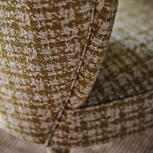 Contemporary Upholstered Occasional Chair Close-up of the textured upholstery on the Contemporary Upholstered Occasional Chair. The fabric combines brown and beige tones, forming a speckled pattern. Detailed stitching lines the edge, and the chair is positioned to show a partial view of both the seat and armrest. A wooden floor lies beneath it.