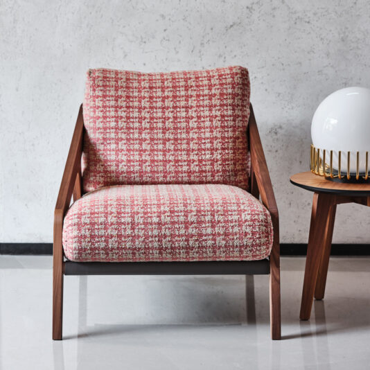 A **Contemporary Walnut Arm Chair** featuring red and white checkered fabric upholstery rests on a glossy floor. The chair showcases wooden armrests and legs. To its right, there's a small round wooden side table topped with a white spherical lamp. The backdrop is a light-textured wall.