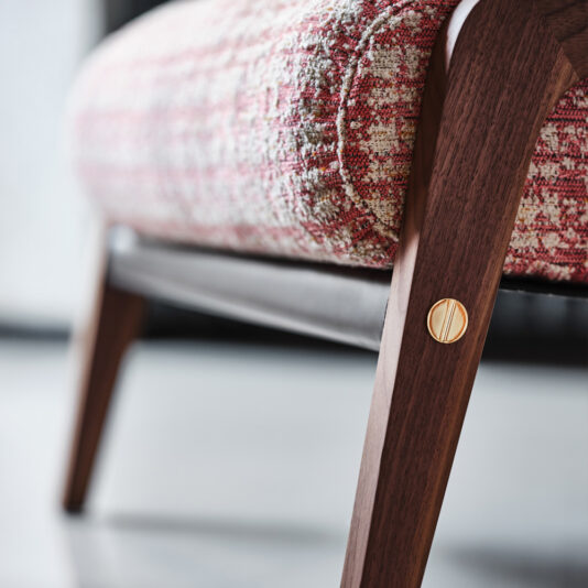 Close-up of a stylish wooden leg from the Contemporary Walnut Arm Chair and Footstool, showcasing sleek design and craftsmanship. The chair features textured fabric with a red and white pattern, and a brass decorative button on the leg adds elegance. A softly blurred background enhances the aesthetic appeal.
