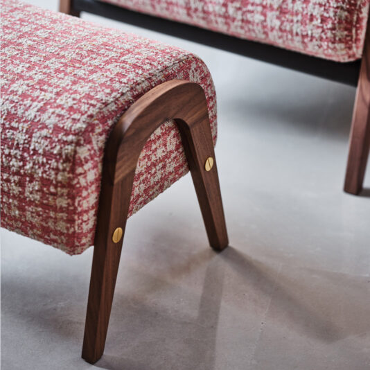 A close-up of the Contemporary Walnut Arm Chair, which boasts a red and white tweed upholstered seat and wooden legs accented with brass hardware, resting on a light grey floor. In the background, part of the matching footstool from the set is partially visible.