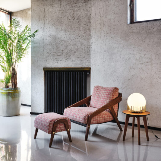 A modern interior features the Contemporary Walnut Arm Chair and Footstool with pink patterned upholstery. A small wooden side table holds a spherical lamp with a unique wooden base. A tall potted plant stands nearby, and a vertical radiator is mounted on the wall.