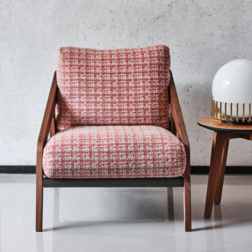Contemporary Walnut Arm Chair and Footstool A stylish armchair from the Contemporary Walnut Arm Chair and Footstool set, featuring a red and white checkered fabric cushion and wooden armrests, sits against a minimalist gray wall. Positioned beside it is the matching footstool, accompanied by a small round side table that holds a white spherical lamp with a gold-colored base.