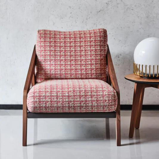 A stylish armchair from the Contemporary Walnut Arm Chair and Footstool set, featuring a red and white checkered fabric cushion and wooden armrests, sits against a minimalist gray wall. Positioned beside it is the matching footstool, accompanied by a small round side table that holds a white spherical lamp with a gold-colored base.