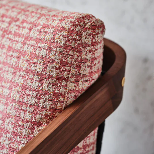 Close-up of the Contemporary Walnut Arm Chair showcasing a textured fabric cushion in red and white hues, framed by a sleek wooden structure with a curved design. The polished walnut features visible grain and a brass accent screw, paired perfectly with its matching footstool.