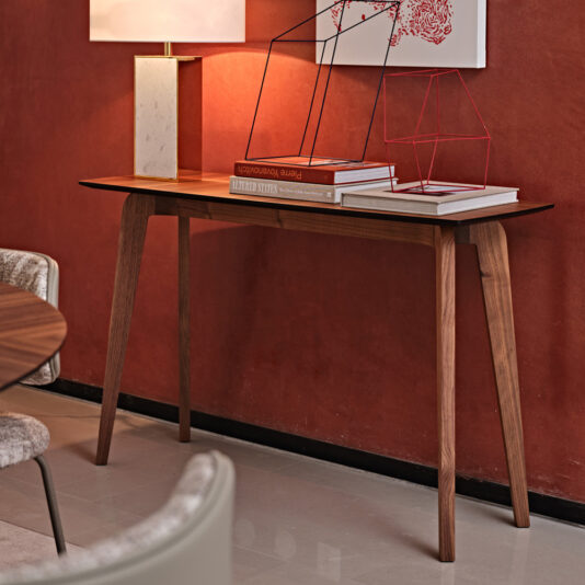 A Contemporary Walnut Console Table with tapered legs is placed against a red wall. On the table are a lamp, two wireframe sculptures, and a few stacked books. The scene is complemented with modern art on the wall and a round chair partially visible in the foreground.