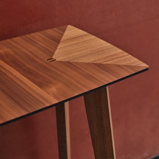 A close-up of the Contemporary Walnut Console Table against a red background. The table showcases a geometric, triangular pattern on its surface, featuring a small circular hole near the center. Its natural finish highlights the wood grain and rich, warm tones of the walnut material.